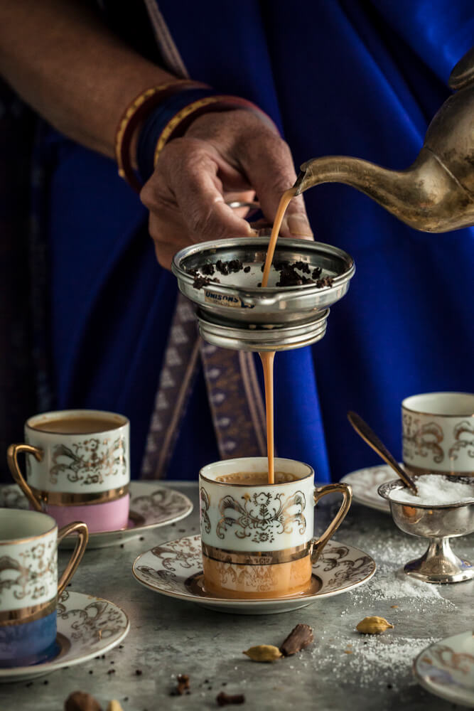 Masala chai being poured through a strainer into floral teacups
